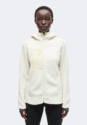 Personne à la peau foncée portant une veste polaire blanche avec une poche poitrine zippée, debout devant un fond clair uni.