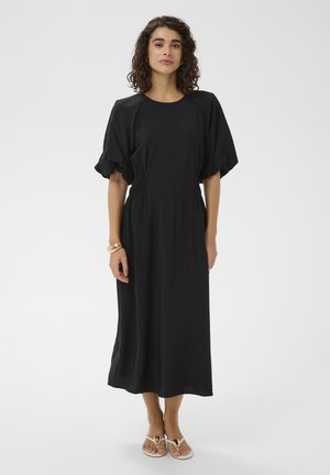 Kobieta o kręconych włosach, ubrana w czarną sukienkę midi z bufkami na rękawach, beżowe sandały oraz złotą bransoletkę, stojąca na jednolitym tle.