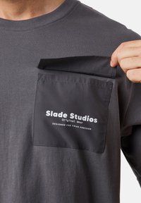 Harmaa t-paita, jossa on kontrastivärinen tummaharmaa tasku valkoisella brändäyksellä. Taskun läppä on osittain nostettu, paljastaen sileän kankaan rakenteen.