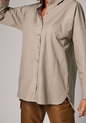 Chemise beige à manches longues boutonnée, portée non rentrée sur un pantalon en cuir marron avec poches cargo, un bras levé.