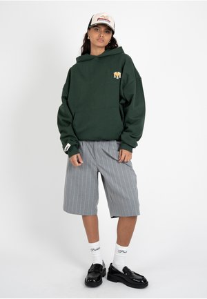 Ung kvinde iført mørkegrøn hættetrøje, grå knælange shorts med striber, hvide sokker, sorte loafers og hvid baseballkasket, poserer foran hvid baggrund.