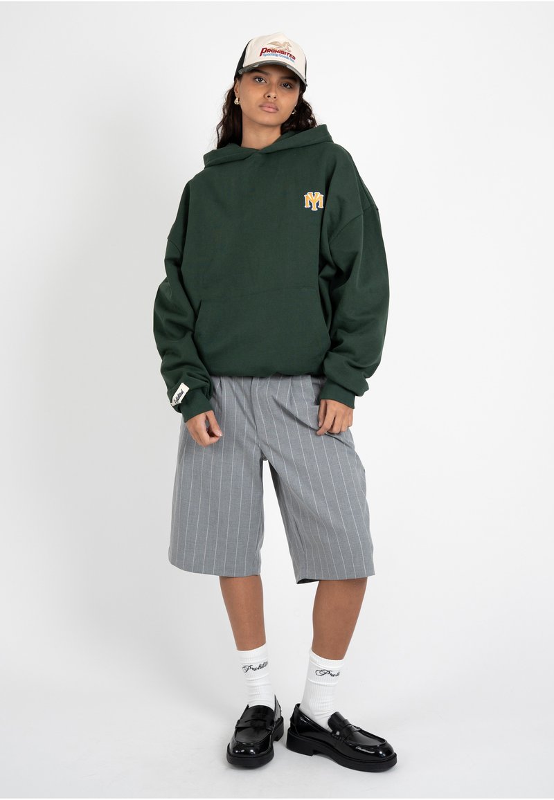 Jeune femme portant un sweat à capuche vert foncé, un short gris à fines rayures jusqu’au genou, des chaussettes blanches, des mocassins noirs et une casquette de baseball blanche, posant sur fond blanc.