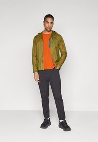Lightweight olive green hooded jacket over an orange knit sweater, paired with dark tapered pants and black athletic shoes.