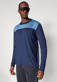 Marineblauwe longsleeve shirt met een lichtblauwe bovenpaneel en logo. Heeft een gladde textuur en een ronde hals, gecombineerd met donkere sportbroek.