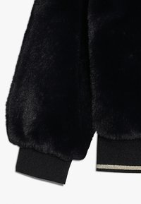 Svart fejkpäls bombarjacka med strukturerad tyg, voluminösa ärmar och ribbade svarta muddar med en kontrasterande metallisk detalj.