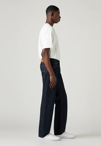 Jeans de mezclilla azul oscuro con un corte recto, con costuras claras, bolsillos traseros y un parche de cuero marrón en la cintura.