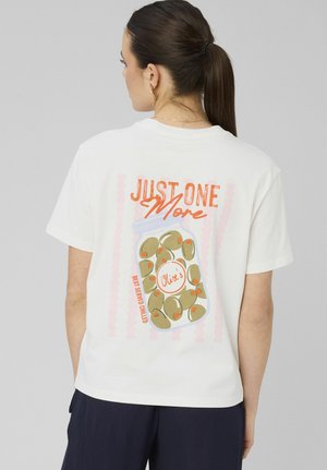 Femme avec queue de cheval foncée portant un T-shirt blanc affichant au dos le texte « Just One More » et un dessin de pot d'olives en orange et vert.