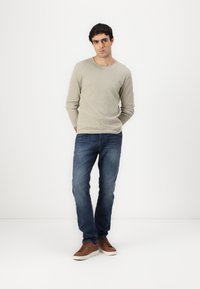 Camisa de punto de manga larga color beige claro, jeans de mezclilla azules y zapatillas de cordones marrones. La camisa tiene un escote redondo y una tela texturizada.