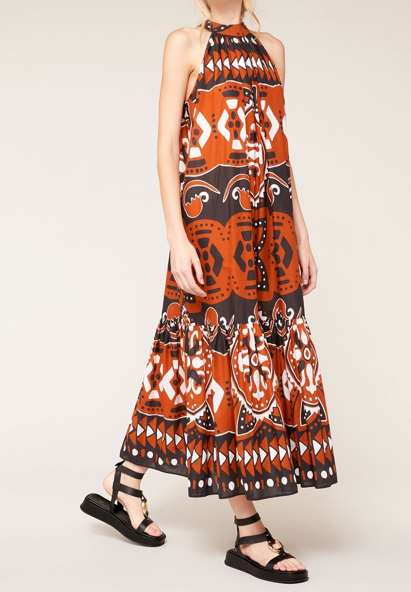 Robe maxi orange et noire avec des motifs tribaux, design à col halter et ourlet froncé. Associée à des sandales noires à lanières.