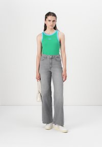 Top verde cu margini albastre, blugi gri cu talie înaltă și adidași albi. Modelul ține o mică poșetă albă. Fundal minimalist.