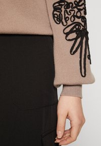 Pull beige en tricot avec détails brodés noirs sur la manche, associé à un bas en tissu foncé doté d'une poche.