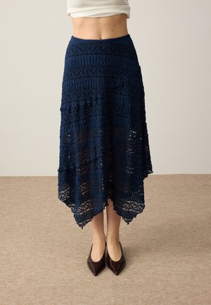 Zużyta granatowa koronkowa spódnica midi z teksturowanymi poziomymi wzorami i asymetrycznym dołem, zestawiona z ciemnymi szpilkami o spiczastym nosku.