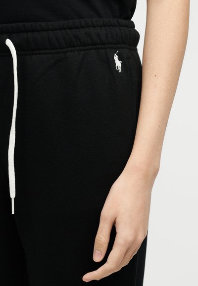 Μαύρο παντελόνι φόρμας με λευκό κορδόνι και λογότυπο Polo Ralph Lauren κοντά στην τσέπη, εμφανίζεται δίπλα σε ένα χαλαρό χέρι και μέρος ενός αντιβραχίου.