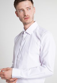 Bela dolga srajca z dolgimi rokavi, narejena iz gladkega bombaža, z najlonskim ovratnikom, sprednjim gumbom in prilagojenim krojem ter kontrastnimi gumbi.