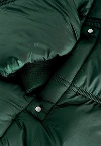 Tamnozeleni puffer kaput s glatkom, sjajnom tkaninom, kviltanim uzorkom, rebrastim manžetama i detaljem na kopčanje s dugmetima.