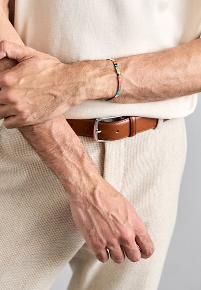 Bracciale multicolore di perline con perline cilindriche rosa, blu e gialle. Indossato su un polso, mostra un mix di motivi. Cintura in pelle marrone visibile.