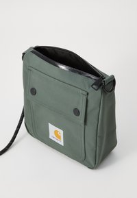 Borsa a tracolla verde in tela con zip superiore, tasca anteriore con patta, hardware nero e patch del logo visibile. Superficie testurizzata, forma compatta.