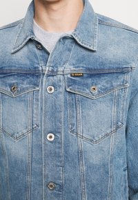 Ljusblå denimjacka med knappstängning, två bröstfickor, silverdetaljer och kontrasterande sömmar. Logotag: "G-STAR."