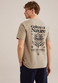 Beige, rövid ujjas póló, fekete grafikai felirattal és virágos mintázattal a hátsó oldalon. Az anyag lágy érzetűnek tűnik, kényelmes szabással.