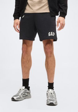 Homme portant un short noir GAP, une chemise beige, une veste noire, des chaussettes montantes noires et des chaussures de course argentées et blanches, debout sur fond blanc.