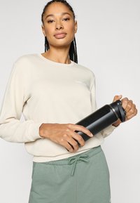 Vannflaske i svart rustfritt stål med sikkert lokk, holdt i en lys beige langermet skjorte og grønne bukser med snor.