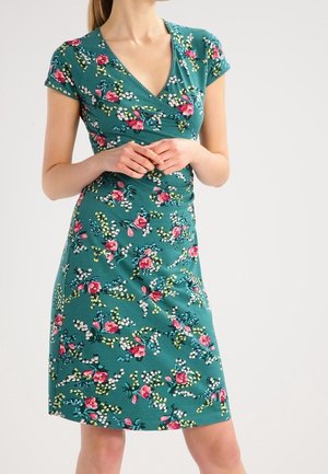 Kobieta ubrana w turkusową sukienkę w kwiaty na krótkie rękawy z różowymi i białymi kwiatami, delikatnie splecione ręce na talii, na jednolitym tle.