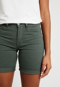 Shorts de mezclilla verdes con bolsillos delanteros, cierre de botón y dobladillo enrollado. La tela parece suave con una textura lisa.