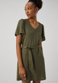 Vestido curto verde-azeitona em tecido leve, com mangas bufantes, decote em V e um cinto do mesmo tecido amarrado na cintura.