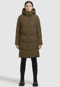 Langer olivgrüner, gepolsterter Mantel mit Kapuze, ausgestattet mit einem Reißverschluss und Druckknopfverschluss, Brust- und Seitentaschen sowie einem gesteppten Design. Schwarze kniehohe Stiefel.