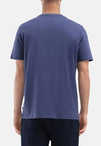 Man in een effen blauwe t-shirt met korte mouwen en donkere broek, gezien vanuit de achterkant tegen een witte achtergrond.