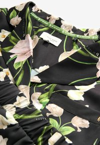 Zwarte stoffen blouse met een bloemenprint van witte en groene bloemen. Bevat een geraffineerd detail op de mouw en een labeltag op de nek.