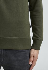 Sudadera de color verde oliva con mangas largas y puños acanalados, que presenta un acabado texturizado. Combinada con unos jeans oscuros grises, mostrando la colocación de las manos.