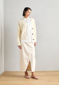 Cardigan crème avec des côtes horizontales, sept boutons et une poche, associé à une jupe maxi crème avec une fente diagonale. Ballerines roses.