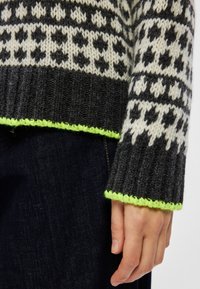 Pulover tricotat cu un model în culori negre și crem, cu mansete cu zimți și un accent verde neon la tiv. Textură din lână sau similară.