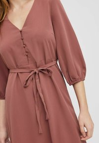 Różowa sukienka z dekoltem w kształcie litery V, bufkami, dopasowanym wcięciem w talii i związanym paskiem. Posiada dekoracyjne guziki z przodu. Miękka tekstura materiału.