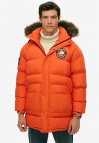 Lys orange pufferjakke med en hætte foret med pels, trykknapper og et patch-logo på brystet; har to frontlommer og en quiltet tekstur.
