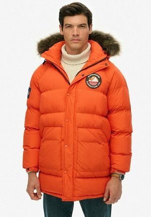 Leuchtend orangefarbene Pufferjacke mit einem mit Pelz gefütterten Kapuze, Druckknöpfen und einem Patch-Logo auf der Brust; verfügt über zwei Fronttaschen und eine gesteppten Textur.