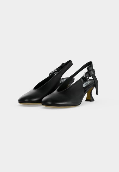 Ζευγάρι μαύρα δερμάτινα πέδιλα slingback με μυτερές μύτες, ασημί λουράκια με αγκράφες και χαμηλά καμπυλωτά τετράγωνα τακούνια σε ανοιχτό φόντο.