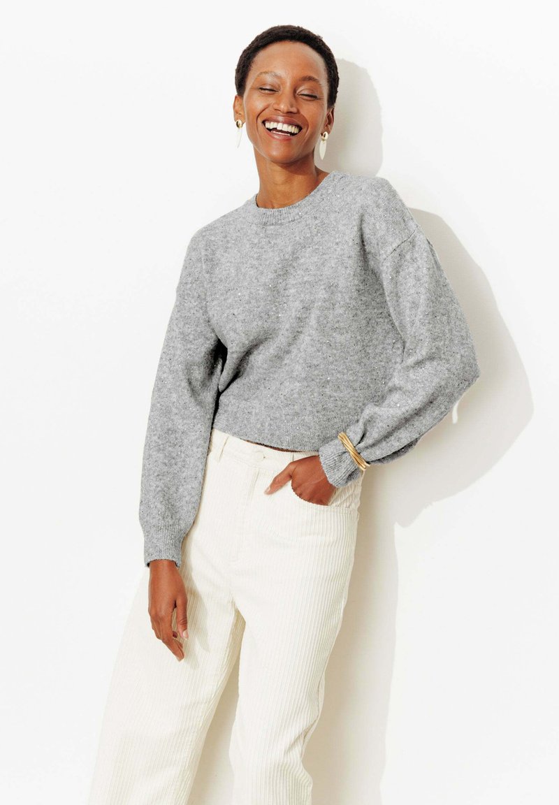 Pull gris en tricot avec une texture côtelée et un col rond, associé à un pantalon taille haute en velours côtelé blanc. Bracelet doré minimaliste visible.