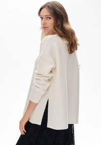 Kobieta z falującymi brązowymi włosami, ubrana w oversize'owy sweter w kolorze kremowym z prążkowanej tkaniny oraz czarną koronkową spódnicę, spoglądająca przez ramię na tle białego tła.