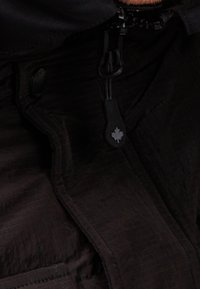 Schwarze Jacke mit strukturiertem Stoff, Reißverschluss und kleinem Ahornblatt-Logo am Reißverschluss. Akzente mit Knopfverschlussdetails.