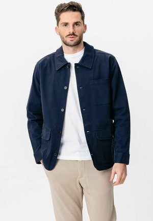 Férfi tengerészkék, gombos dzsekiben, fehér pólóval és bézs nadrággal, egy kezét zsebében tartva, egyszínű háttér előtt áll.