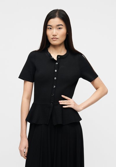 Γυναίκα που φοράει μαύρο κοντομάνικο μπλουζάκι με κουμπιά και ζωνάκι στο κάτω μέρος και μαύρη φούστα που ταιριάζει, στέκεται με το ένα χέρι στη μέση.