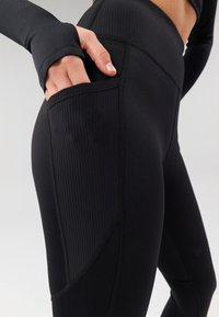 Leggings de sport noirs en tissu lisse, dotés d'une poche latérale côtelée et de coutures plates pour une durabilité et un confort accrus.