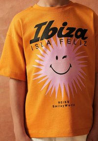 Oranžinė medvilninė marškinė su juodu „Ibiza“ tekstu ir šypseną turinčiu rožiniu saulės grafiku, papildyta prekės ženklu „REISS SmileyWorld“ apačioje.