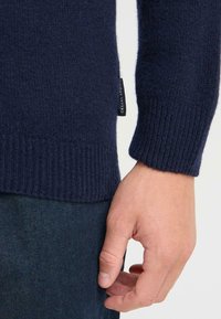 Marine gebreide trui met geribde manchetten, gemaakt van zachte stof. Bevat een label met 'CASUAL' branding aan de zijkant. Hand geplaatst op de taille.