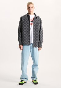 Zwart-wit geblokte overhemd, witte graphic T-shirt, lichtblauwe jeans en zwarte sneakers met neon groene en roze accenten.