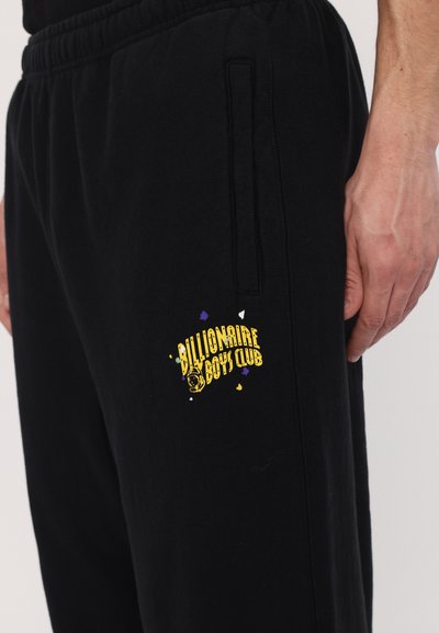 Pantalons de survêtement noirs en tissu doux, avec un logo jaune et violet sur la cuisse gauche, une taille élastique et des poches latérales.