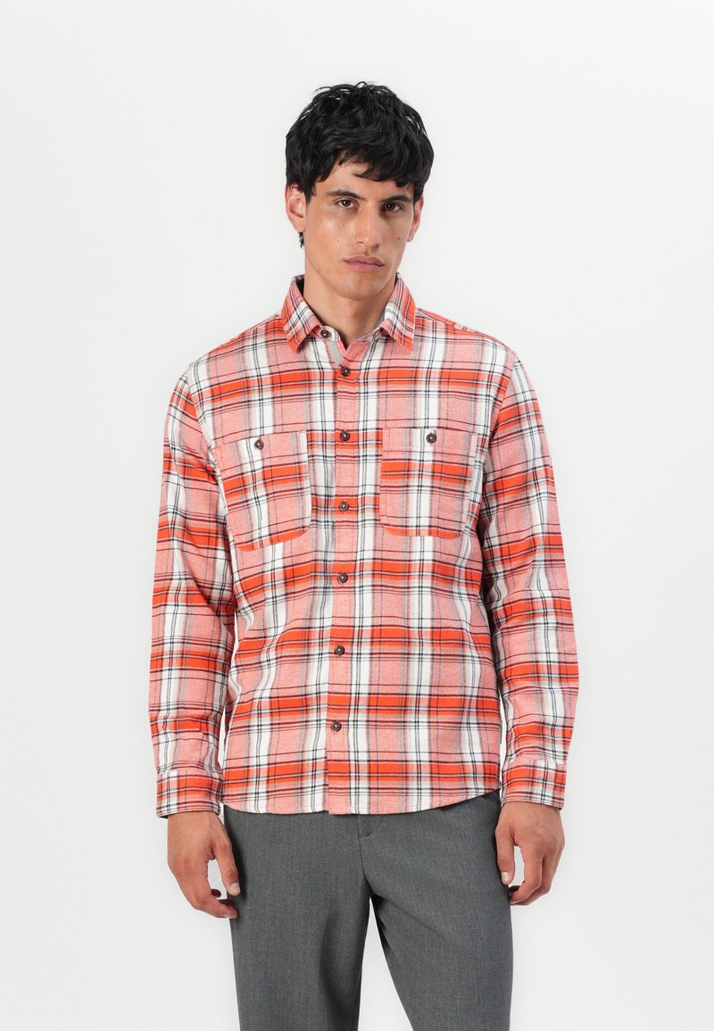 Camicia in flanella a quadri rossi e bianchi con maniche lunghe, due taschini sul petto, chiusura con bottoni e dettagli neri nel motivo. Indossata con pantaloni grigi.