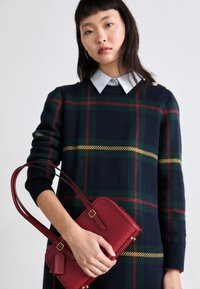 Mörkblå rutig tröja med gröna, gula och röda ränder, med en krage över en ljusblå skjorta; modellen håller en röd handväska med gulddetaljer.